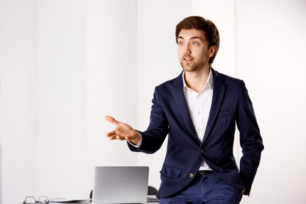 Handsome confused confident young businessman talking near the office desk. Isolated on white background.